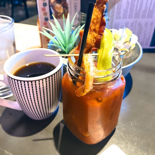 A Bloody Mary in a mason jar with bacon, shrimp, and pepper, next to a cup of coffee.