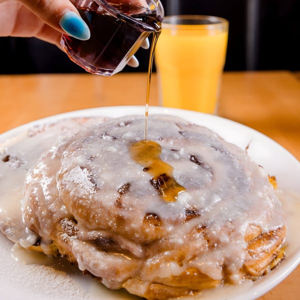 Maple syrup being poured over a large, frosted cinnamon roll with powdered sugar, next to orange juice.
