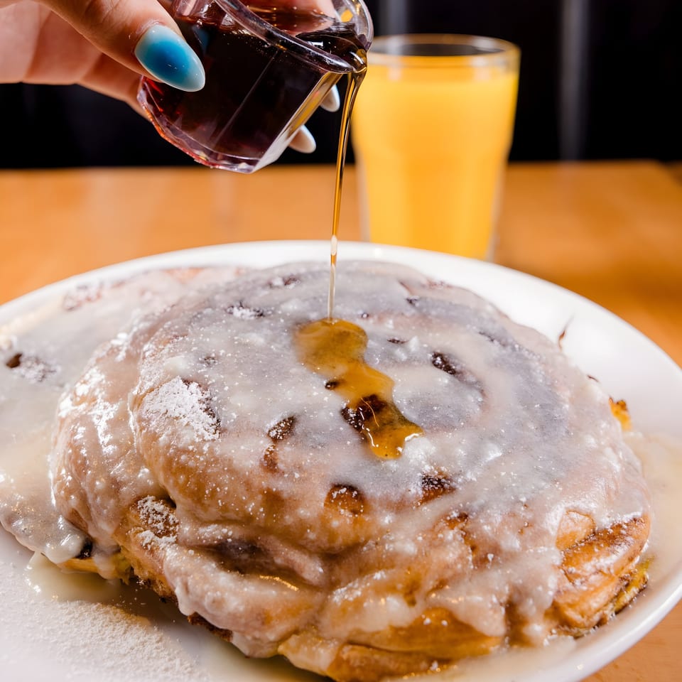 Maple syrup being poured over a large, frosted cinnamon roll with powdered sugar, next to orange juice.