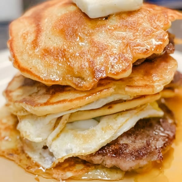 A stack of pancakes, eggs, and sausage patties drizzled with syrup and topped with butter.