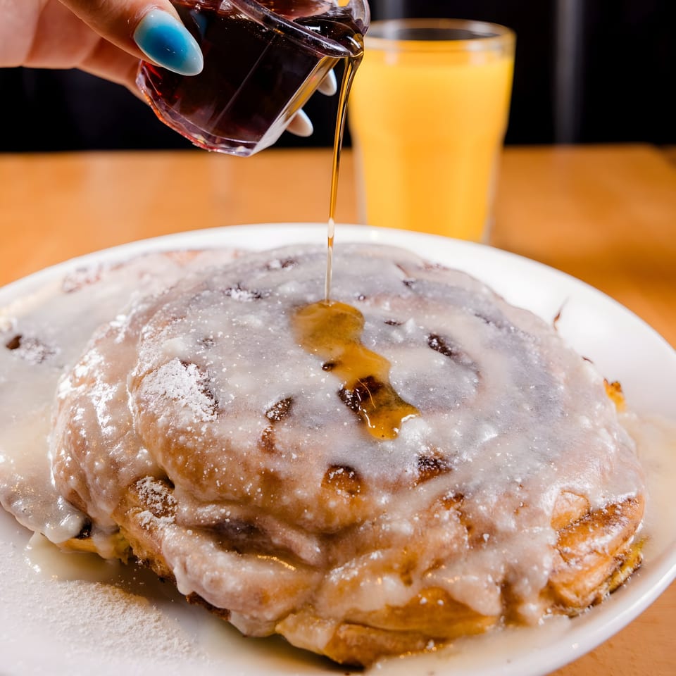 Maple syrup being poured over a large cinnamon roll with white icing and powdered sugar.