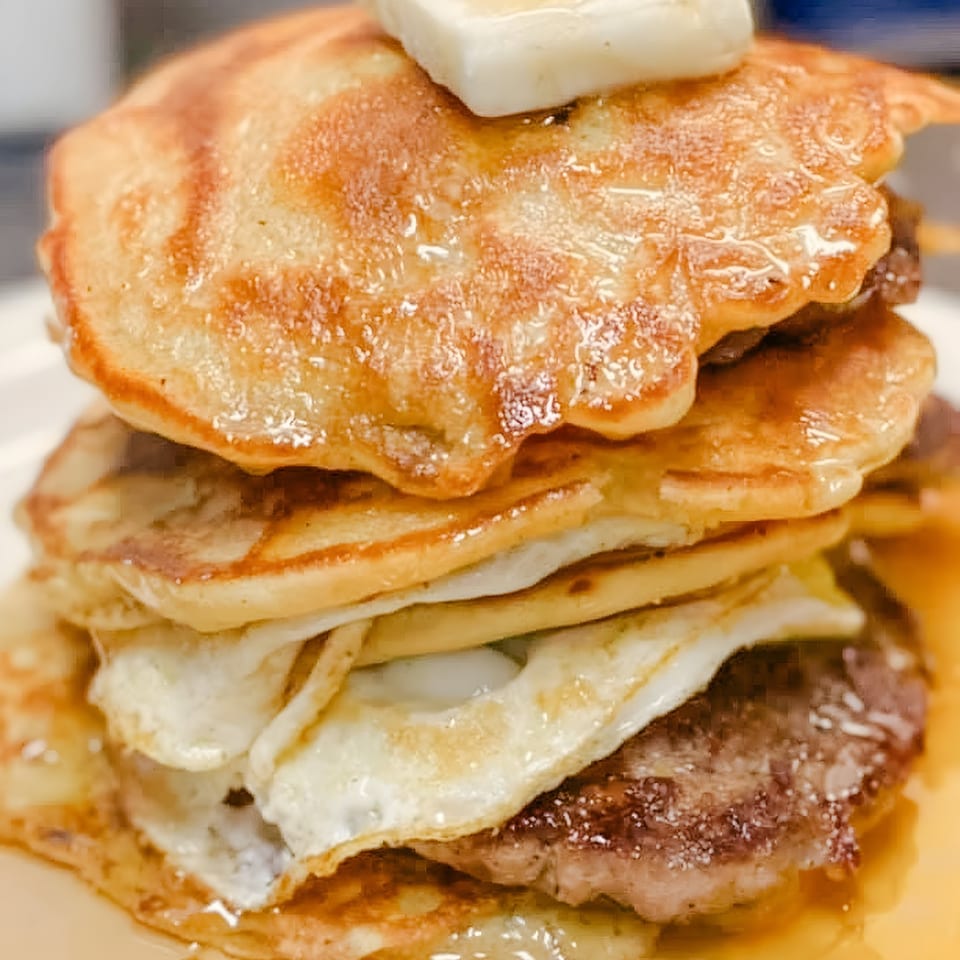 A stack of pancakes, fried eggs, and sausage patties, drizzled with syrup and topped with butter.