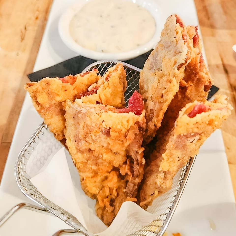 Crispy fried bacon strips in a metal basket, with a side of creamy white dipping sauce.