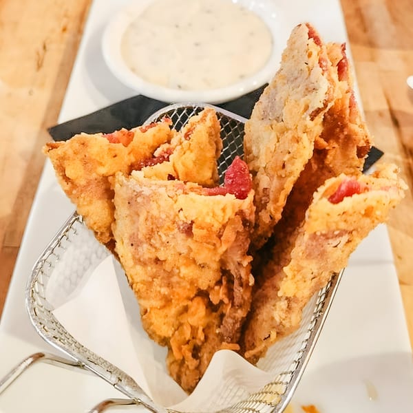 Crispy fried bacon strips in a metal basket, with a side of creamy white dipping sauce.