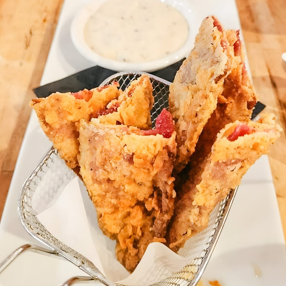 Crispy fried bacon strips in a metal basket, with a side of creamy white dipping sauce.