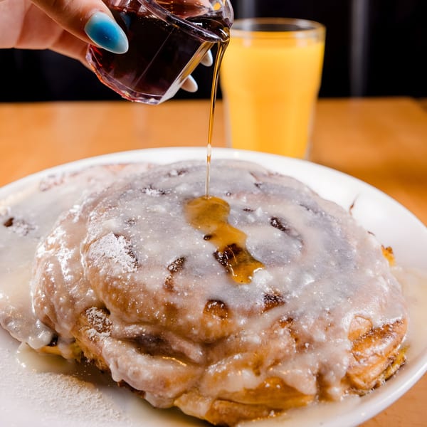 Maple syrup being poured over a large, icing-covered cinnamon roll pancake at Brunch Snob.