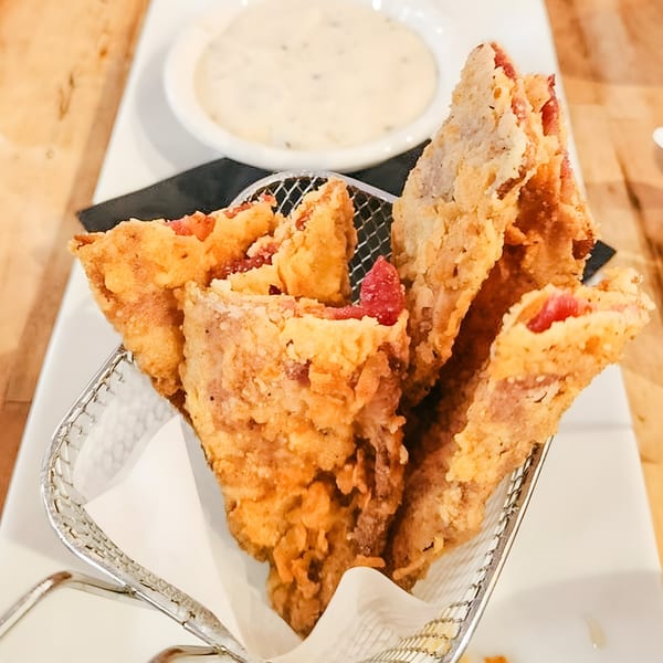 Golden-fried bacon strips in a metal basket, with a side of creamy dipping sauce.