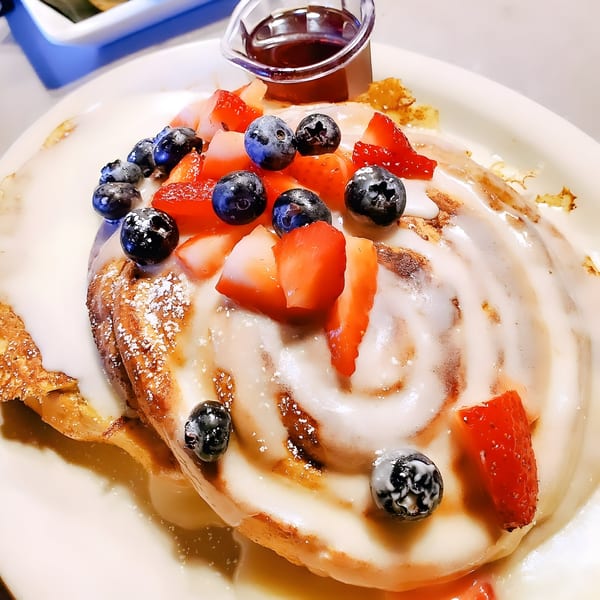 A large cinnamon roll pancake with white icing, fresh blueberries, strawberries, and a side of syrup.